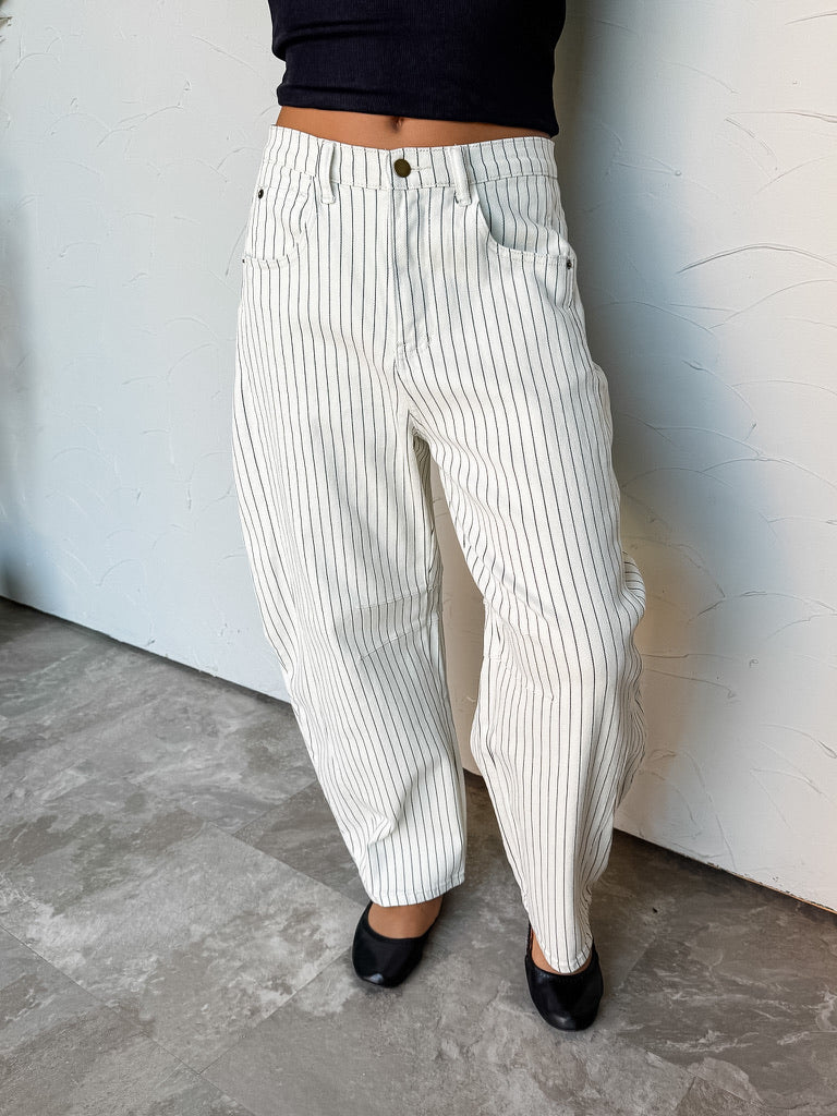 A photograph from the waist down of a model wearing high-waisted white pinstriped barrel jeans against a textured white wall. The pants are crafted from a 98% cotton and 2% spandex blend, featuring black vertical pinstripes, belt loops, five-pocket construction, and a curved, wide barrel-leg shape, paired with a black crop top and black ballet flats.
