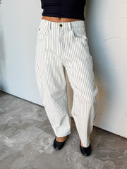 A photograph from the waist down of a model wearing high-waisted white pinstriped barrel jeans against a textured white wall. The pants are crafted from a 98% cotton and 2% spandex blend, featuring black vertical pinstripes, belt loops, five-pocket construction, and a curved, wide barrel-leg shape, paired with a black crop top and black ballet flats.