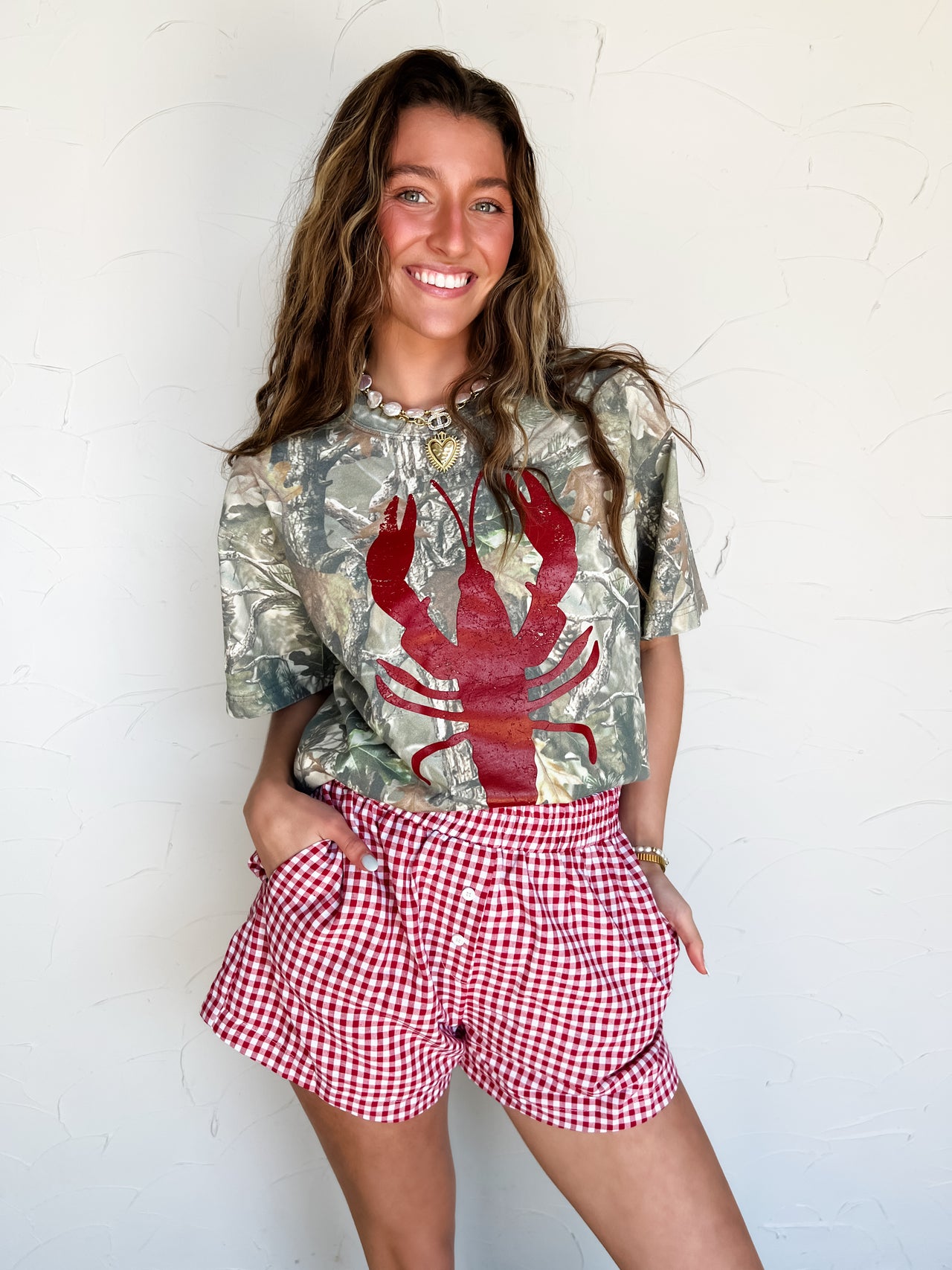 Woman wearing a camouflage shirt with a red lobster design and red checkered shorts against a white wall.