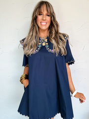 Woman wearing a navy dress with embroidery on the large collar against a white brick wall.