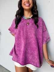 A close-up photograph from the chin down of a smiling brunette woman posing against a textured white wall. She is wearing an oversized crewneck t-shirt made from a 100% cotton fabric with a purple mineral-wash finish, featuring dropped shoulders, short cuffed sleeves, and a raw-edge split hemline.