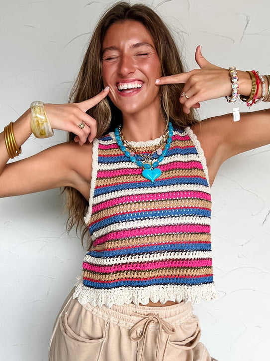 Woman wearing a colorful striped crochet tank top and beige pants against a white wall.
