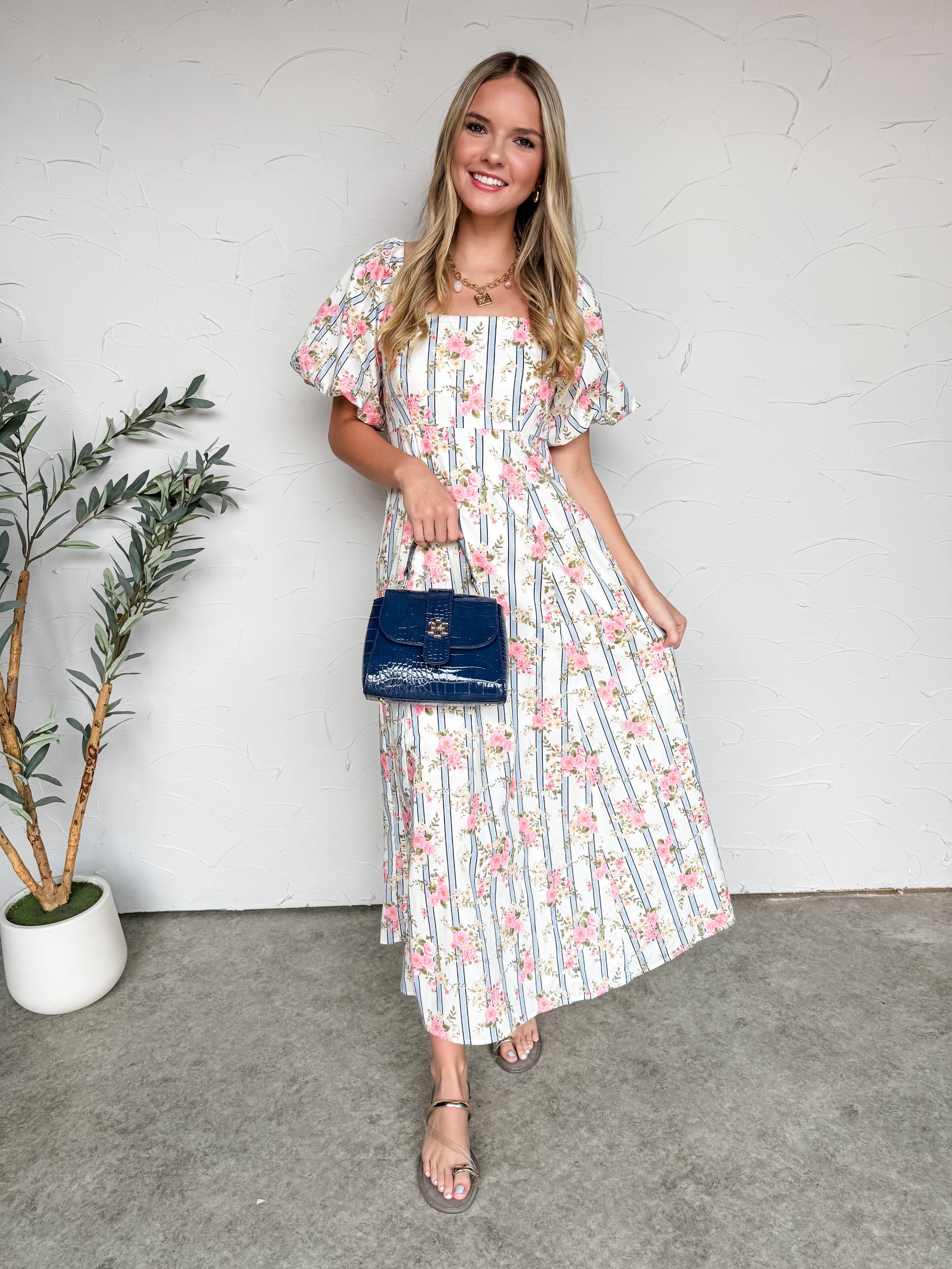 A full-length photo of a smiling blonde woman standing against a textured white wall, showcasing a vintage-inspired maxi dress. The dress features a square neckline, dramatic short puff sleeves, and a fitted high-waisted bodice that flows into a long, breezy A-line skirt. The fabric is white with a pattern of thin, vertical light blue stripes overlaid with clusters of pink flowers and green leaves. 