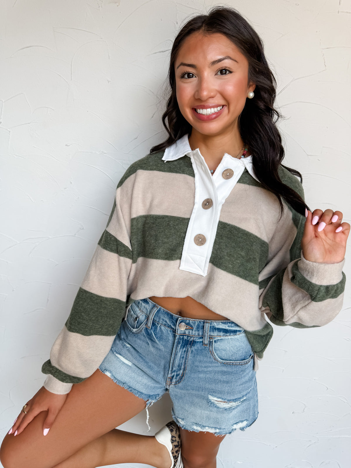 A waist-up photograph of a smiling woman with long dark wavy hair touching a section of her hair while posing with one leg raised against a textured white wall. She is wearing a long-sleeved cropped fleece polo sweatshirt made from an 82% polyester, 15% rayon, and 3% spandex blend, featuring broad olive green and beige horizontal stripes, a contrasting white collar and button placket with large round brown buttons, paired with high-waisted distressed light-wash denim shorts, styled with pearl stud earrings 