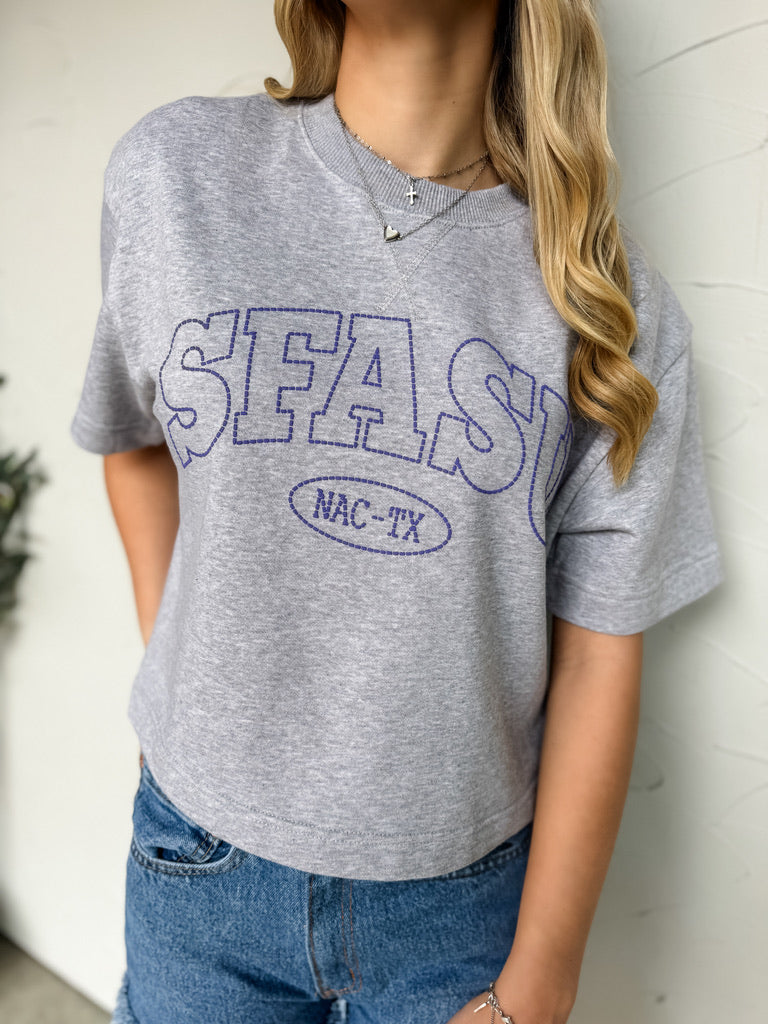 Person wearing a gray t-shirt with 'SFASU' printed on it against a white wall.