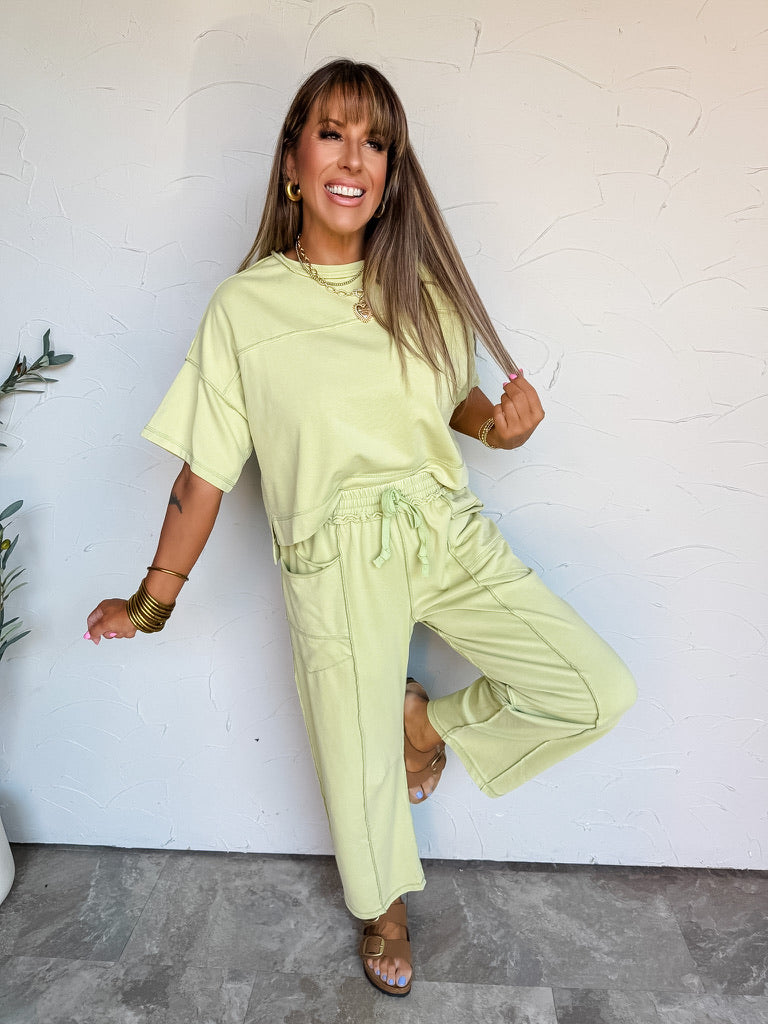 A full-length photograph of a smiling woman with straight light-brown hair and front bangs holding a strand of her hair and posing on one leg against a textured white wall. She is wearing a matching two-piece lime green lounge set featuring a relaxed short-sleeved crewneck top with exposed seam detailing, paired with matching straight-leg pants with an elastic drawstring waist and side pockets, styled with brown slide sandals, stacked gold bangles, gold hoop earrings, and a gold chain heart pendant necklace