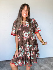 Model wearing the Keep Going Floral Scallop Trim Dress by Entro in 100% polyester, featuring pink and cream flowers with green leaves on a brown background, allover eyelet-style cutouts, a stand collar with a split neckline, short flutter sleeves, and a flared ruffle hem with contrasting rust-red scalloped trim along the sleeves and hem.