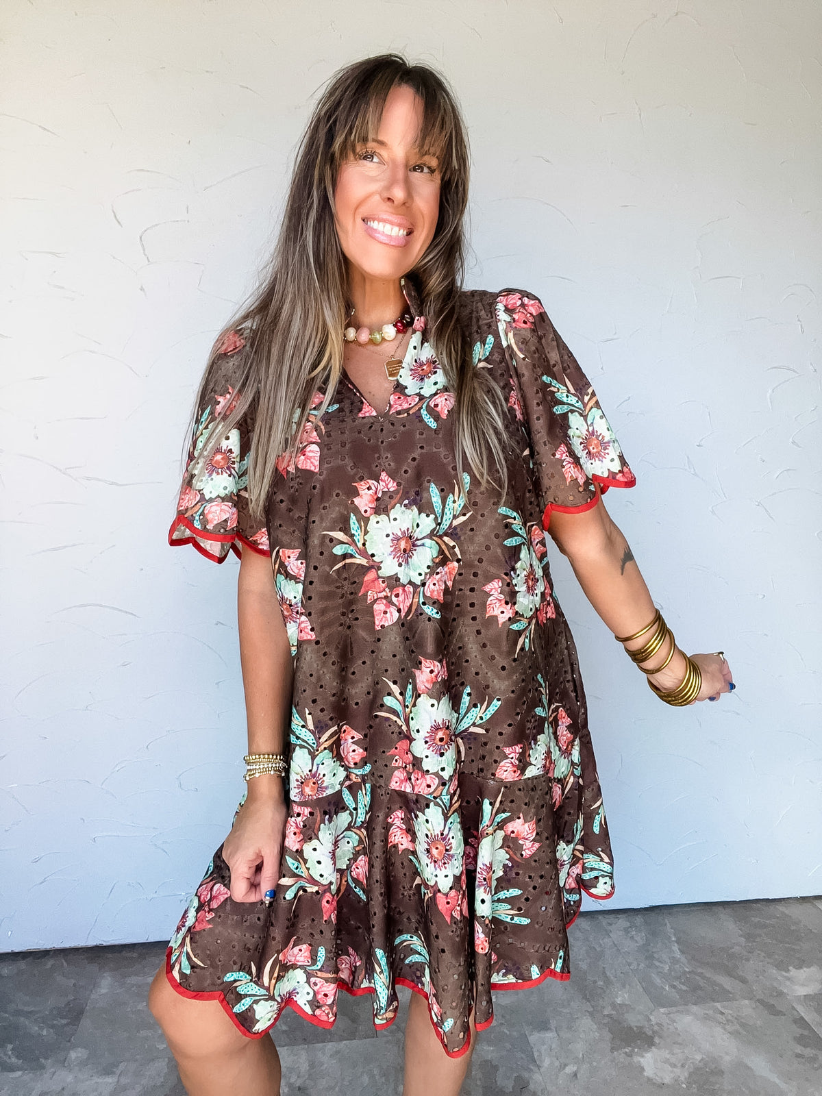 Model wearing the Keep Going Floral Scallop Trim Dress by Entro in 100% polyester, featuring pink and cream flowers with green leaves on a brown background, allover eyelet-style cutouts, a stand collar with a split neckline, short flutter sleeves, and a flared ruffle hem with contrasting rust-red scalloped trim along the sleeves and hem.