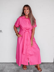 A woman modeling a bright pink short-sleeve shirt dress with a tie waist, standing against a white wall.