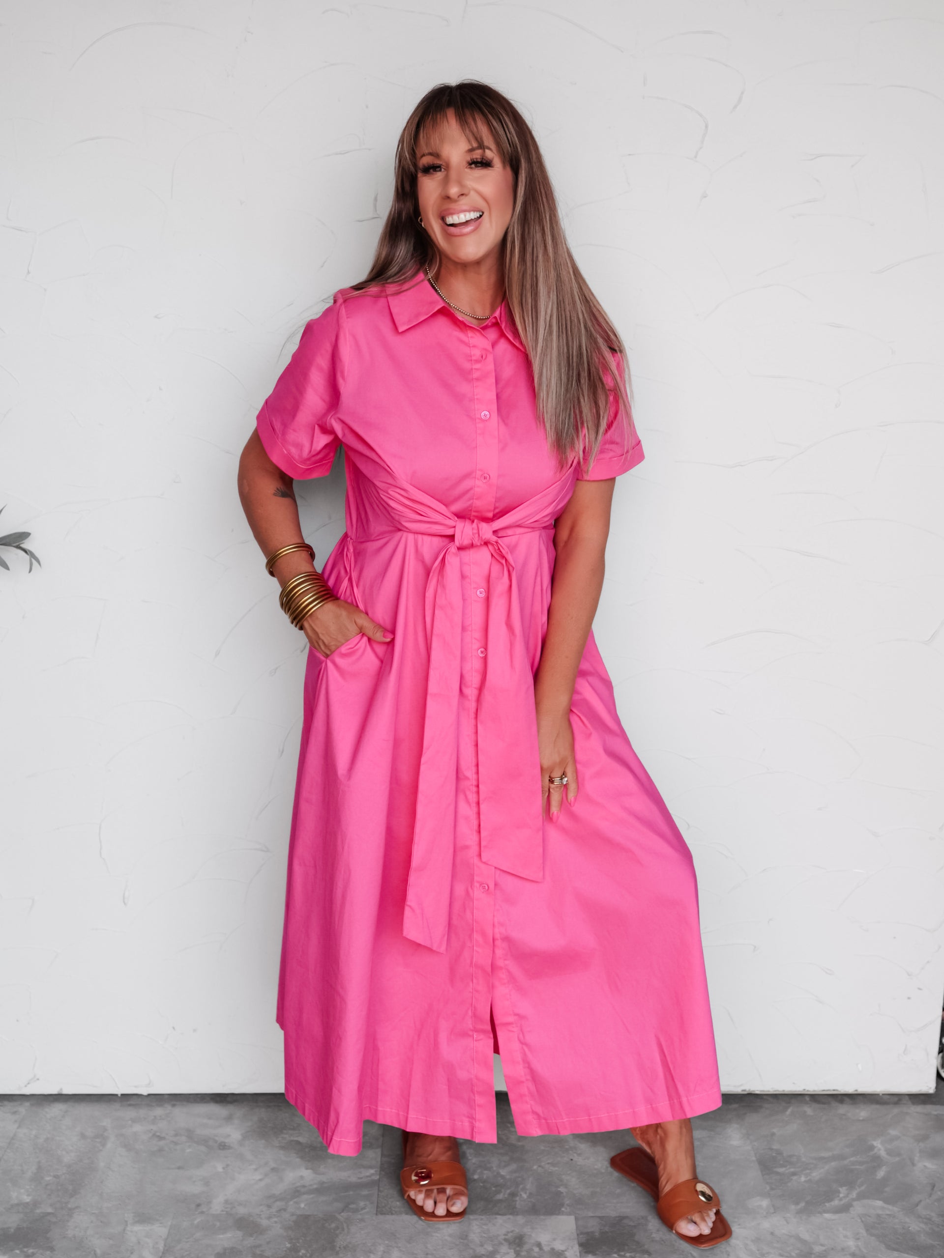A woman modeling a bright pink short-sleeve shirt dress with a tie waist, standing against a white wall.