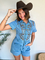 Woman wearing a denim romper with embroidery and a brown hat indoors.