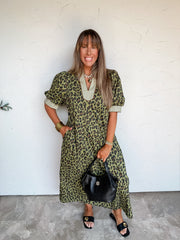 Lesly modeling the Entro Talk of the Town Leopard Print Midi Dress in size Large, made from 100% cotton with olive-toned leopard print, contrasting light patterned trim at the split neckline and wide cuffs, short puff sleeves, and side pockets, styled with black slide sandals, a black handbag, and gold-tone bracelets.