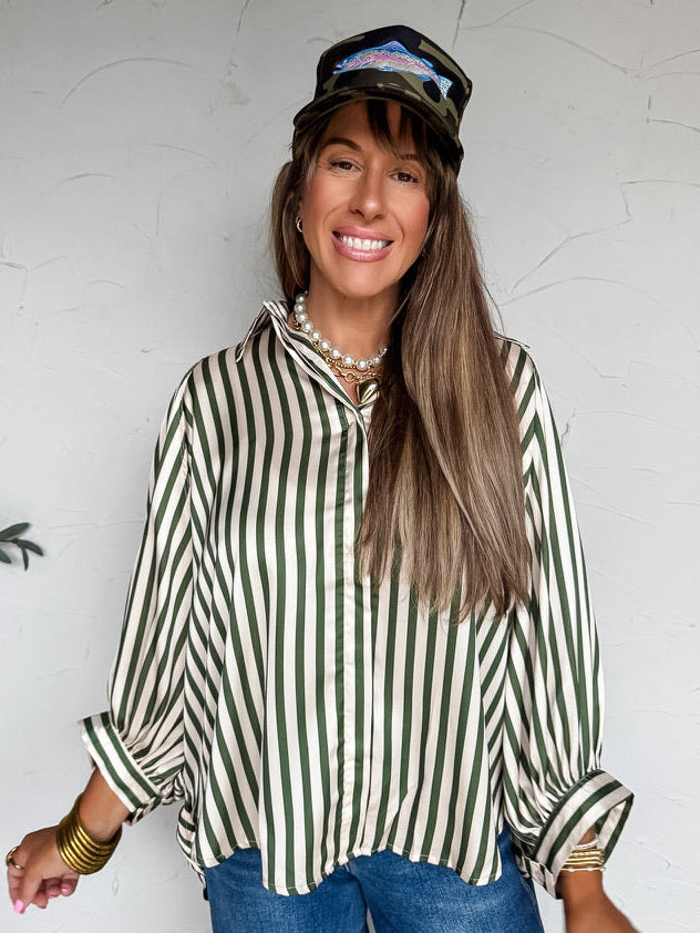 A three-quarter length photograph of a smiling woman with long brown hair and bangs posing against a textured white wall. She is wearing a long-sleeved satin button-down blouse featuring vertical olive green and cream stripes made from 100% polyester, with a pointed collar, relaxed drop-shoulder long sleeves with wide buttoned cuffs, front-tucked into medium-wash blue denim jeans, styled with a camo trucker hat featuring a fish graphic, a layered pearl and gold heart pendant necklace, and stacked gold bangl
