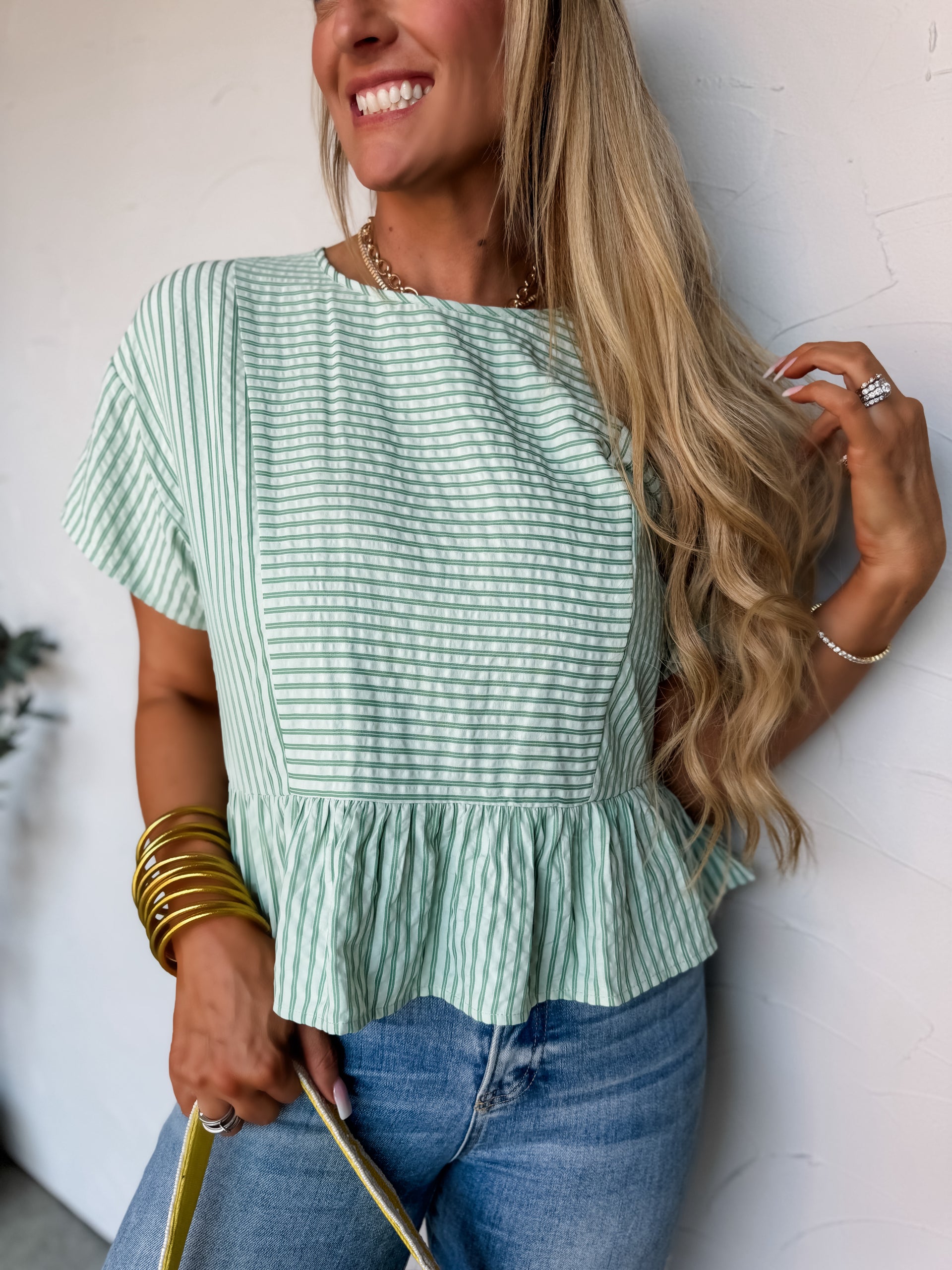A smiling blonde woman leaning against a textured off-white wall, wearing a green and white striped short-sleeve blouse. The top is crafted from a textured 65% polyester and 35% cotton blend seersucker fabric and features a unique combination of horizontal stripes on the front panel and vertical stripes on the sides and peplum hemline. It is designed with a simple crew neckline and loose, short extended sleeves. She is wearing the top with medium-wash blue denim jeans and is accessorized with a thick stack 