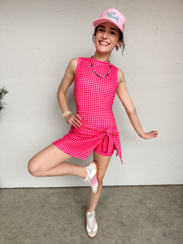 Woman wearing a pink checkered dress and cap posing against a white wall.