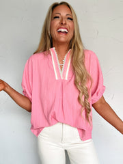 Woman wearing a pink blouse with a v-neck with white details and white pants against a white background