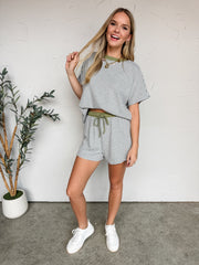 A three-quarter length photo of a smiling blonde woman looking off-camera against a textured white wall. She is wearing a matching grey and white fine-striped lounge set featuring a short-sleeved top with an olive green crew neckline. She playfully lifts the front of her loose shirt with both hands to showcase the contrasting olive green elastic waistband and functional drawstring tie of the matching high-waisted shorts. 