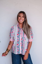 A portrait photograph focusing from the waist up of a smiling woman with straight blonde hair and bangs posing against a textured white wall. She is wearing a short-sleeved button-down blouse featuring vertical stripes of pink, navy, and red floral motifs on a white background, made from 100% cotton fabric, designed with a pink ruffled stand collar, a ruffle-trimmed front placket, and pink striped sleeve cuffs, styled with medium-wash jeans with front center seams and gold bangles.