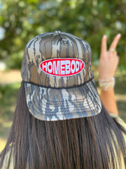 A person shown from behind wearing a camouflage trucker hat with a red and white 'HOMEBODY' patch on the front, with a blurred outdoor background of green foliage.