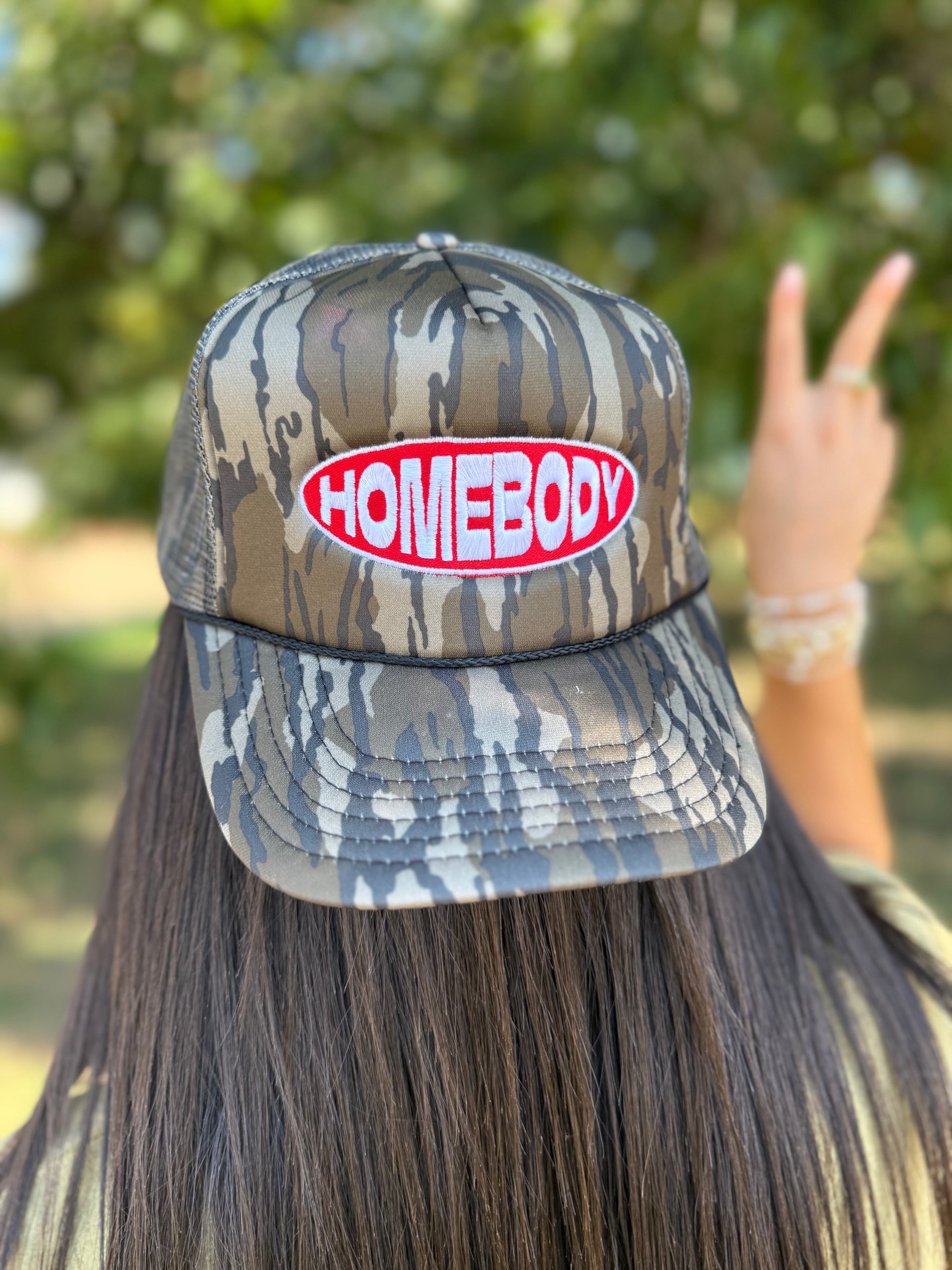 A person shown from behind wearing a camouflage trucker hat with a red and white 'HOMEBODY' patch on the front, with a blurred outdoor background of green foliage.