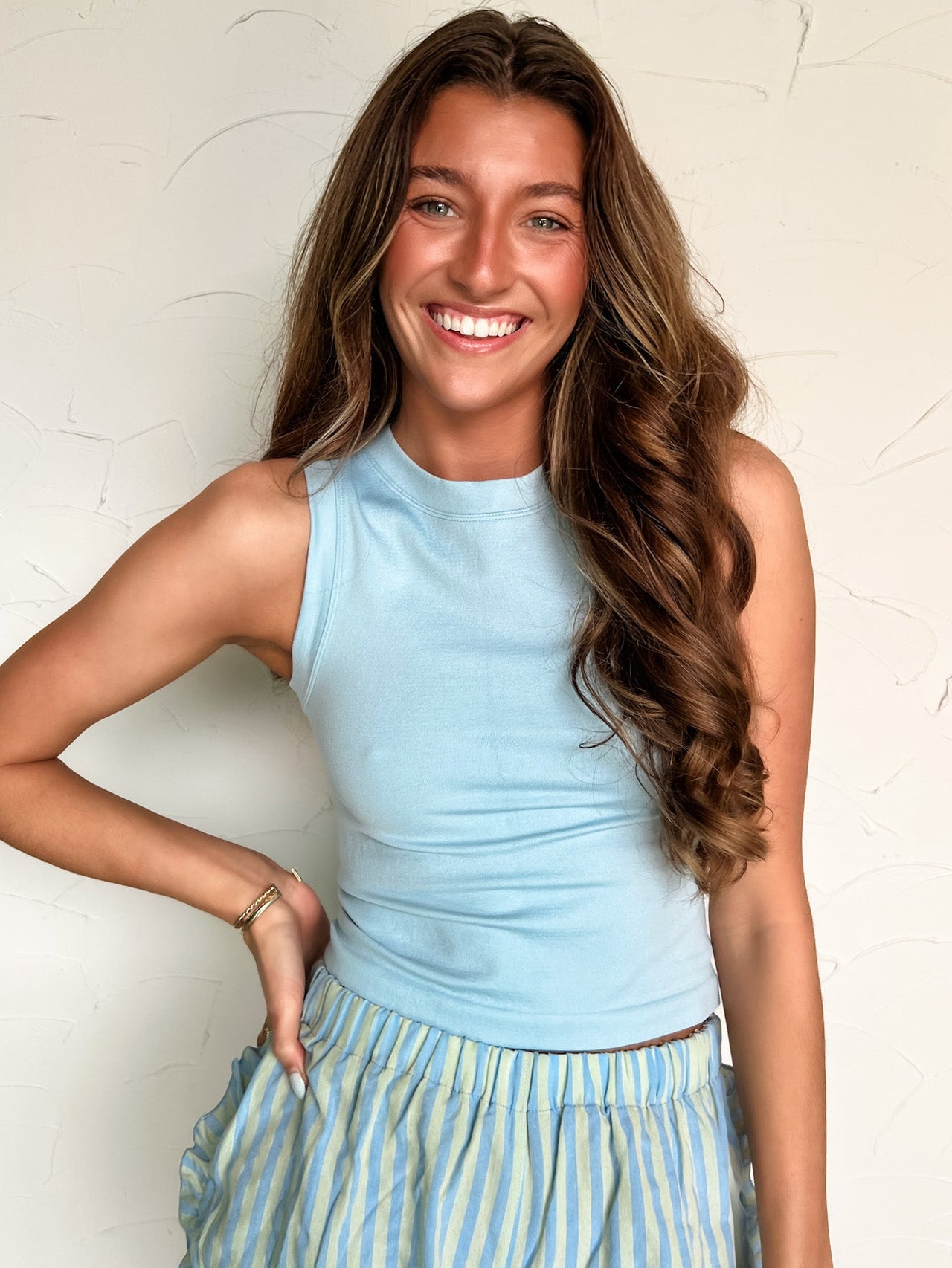 Woman wearing a light blue sleeveless top and striped skirt against a white wall.