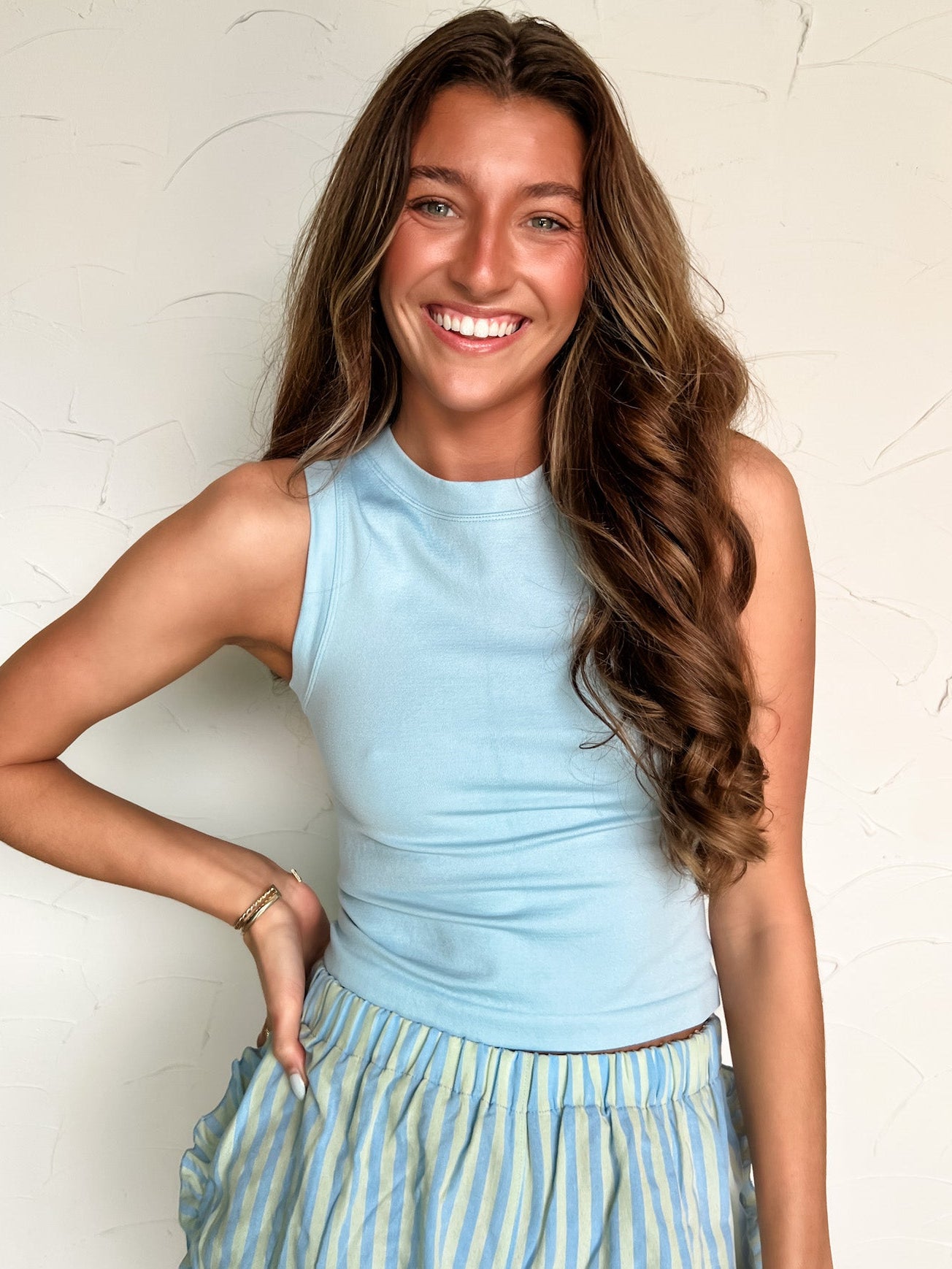Woman wearing a light blue sleeveless top and striped skirt against a white wall.