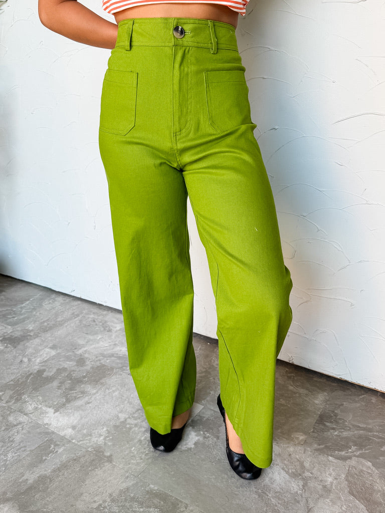 A photograph from the waist down of a model wearing high-waisted lime green wide-leg pants against a textured white wall. The pants are crafted from a 97% cotton and 3% spandex blend, featuring belt loops, a single tortoise-shell button front closure, square front patch pockets, and a full-length wide-leg cut, paired with an orange striped crop top and black ballet flats.