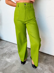 A photograph from the waist down of a model wearing high-waisted lime green wide-leg pants against a textured white wall. The pants are crafted from a 97% cotton and 3% spandex blend, featuring belt loops, a single tortoise-shell button front closure, square front patch pockets, and a full-length wide-leg cut, paired with an orange striped crop top and black ballet flats.