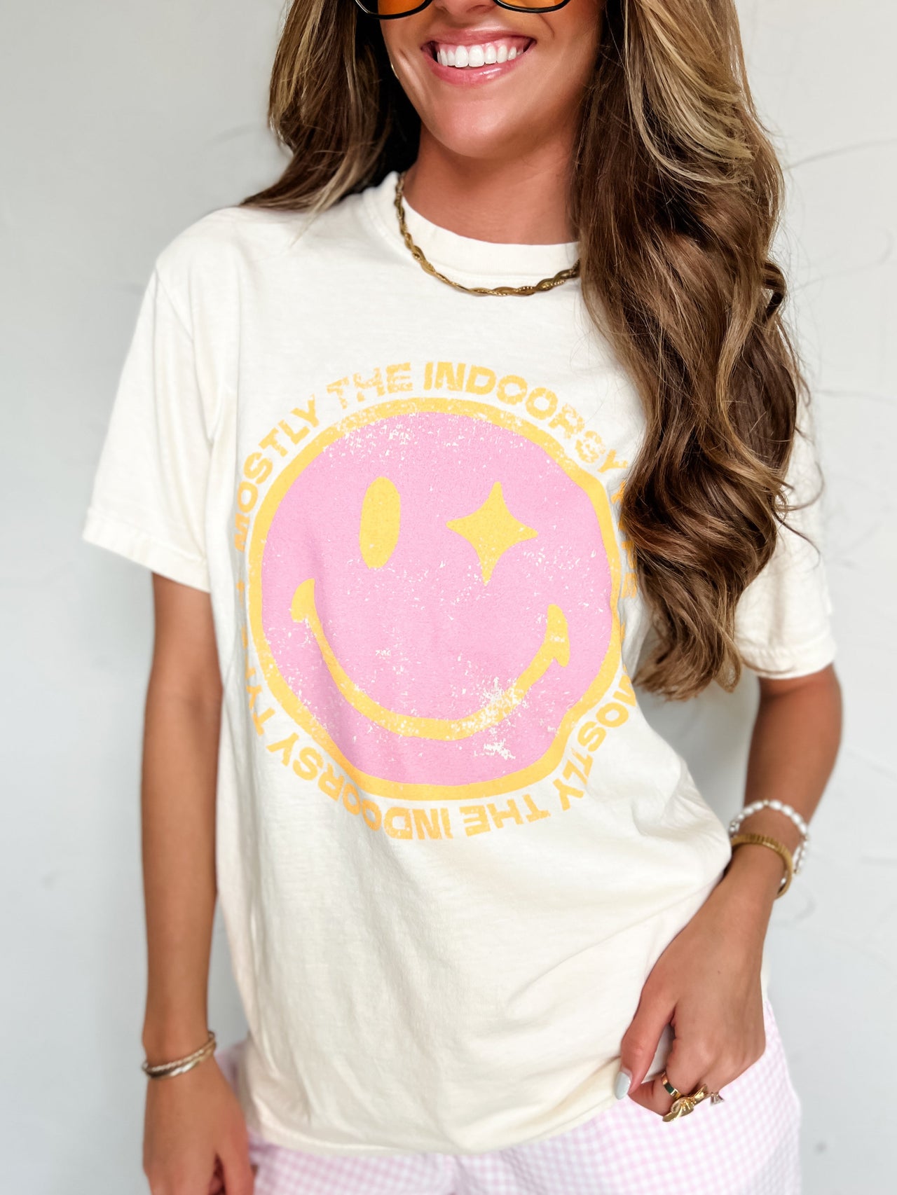 Woman wearing a white t-shirt with a pink smiley face design and sunglasses against a plain background