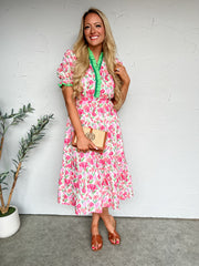Woman in a pink floral dress holding a clutch against a white wall.
