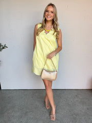 Woman wearing a yellow dress holding a beige handbag against a white wall.