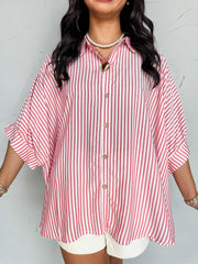 A close-up photograph from the chin down of a smiling brunette woman posing against a textured white wall. She is wearing an oversized button-down shirt made from a lightweight 100% rayon fabric, featuring a red and white vertical stripe pattern, dropped shoulders, and short cuffed sleeves.