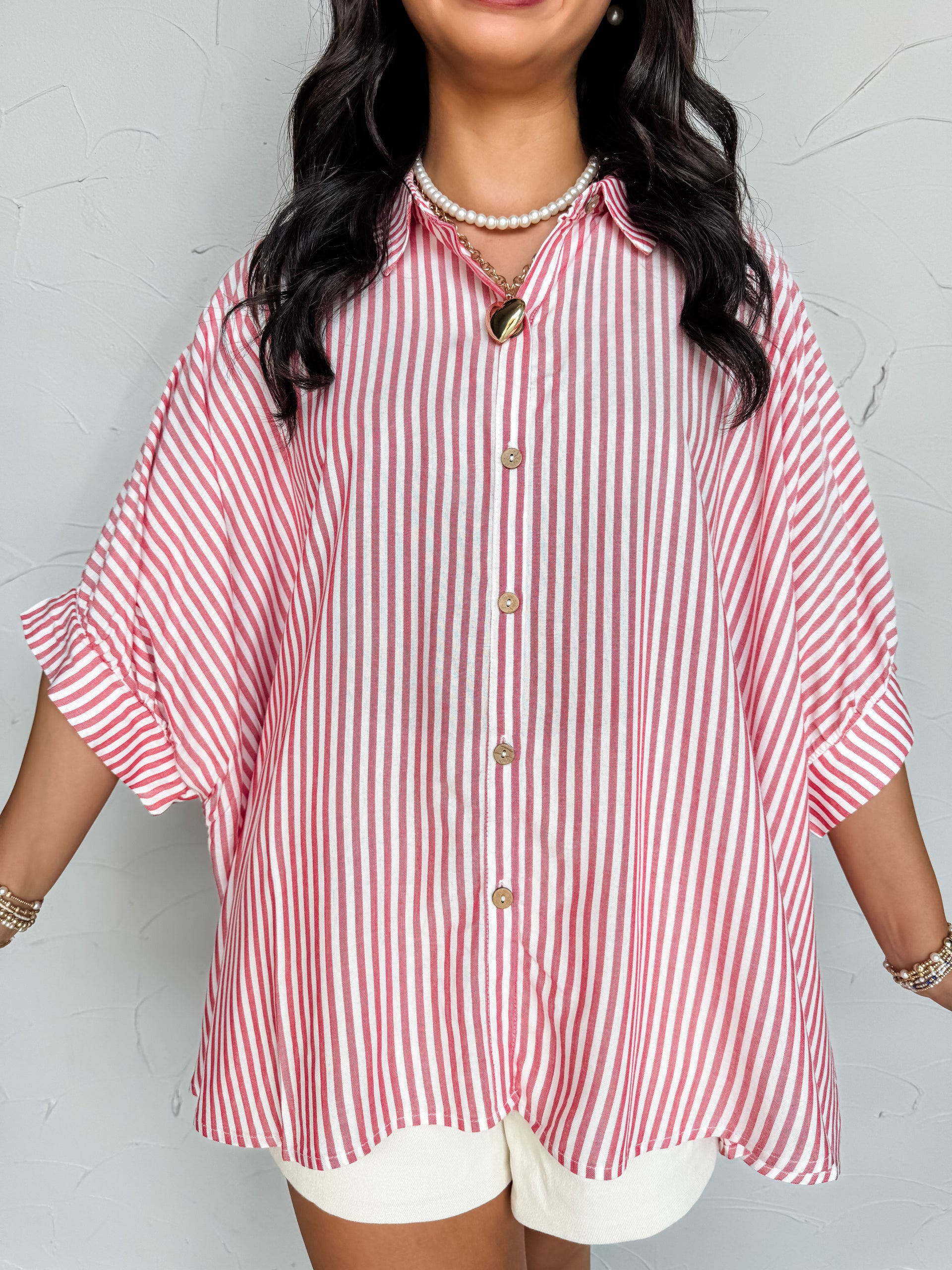 A close-up photograph from the chin down of a smiling brunette woman posing against a textured white wall. She is wearing an oversized button-down shirt made from a lightweight 100% rayon fabric, featuring a red and white vertical stripe pattern, dropped shoulders, and short cuffed sleeves.