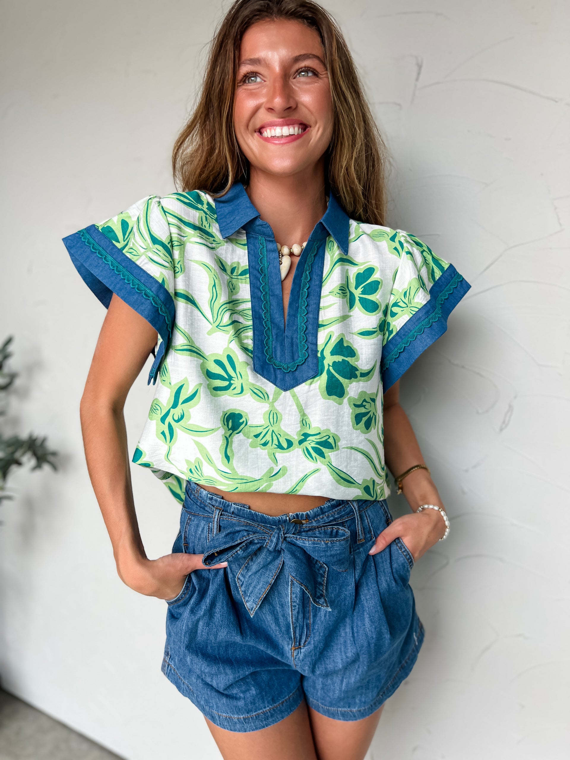 Woman wearing a green and white patterned top with blue accents and denim shorts against a white wall.