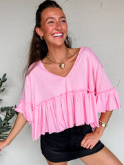 Woman wearing a pink blouse with ruffled sleeves against a white wall.