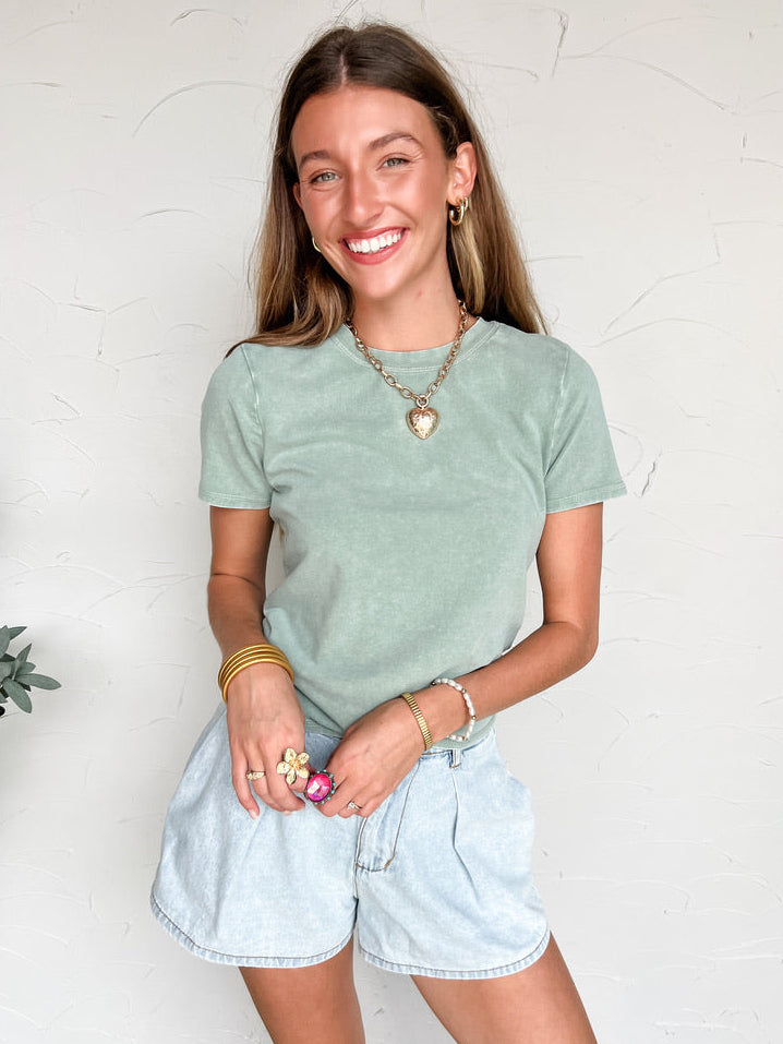 Woman wearing a green t-shirt and light blue denim shorts against a white wall.