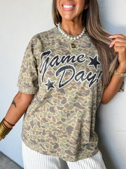 A woman wearing a short-sleeve camouflage graphic t-shirt with 'Game Day!' printed in black glitter-style lettering with star accents. She is accessorized with a pearl necklace, gold bangles, and a gold pendant, posing against a light gray wall.