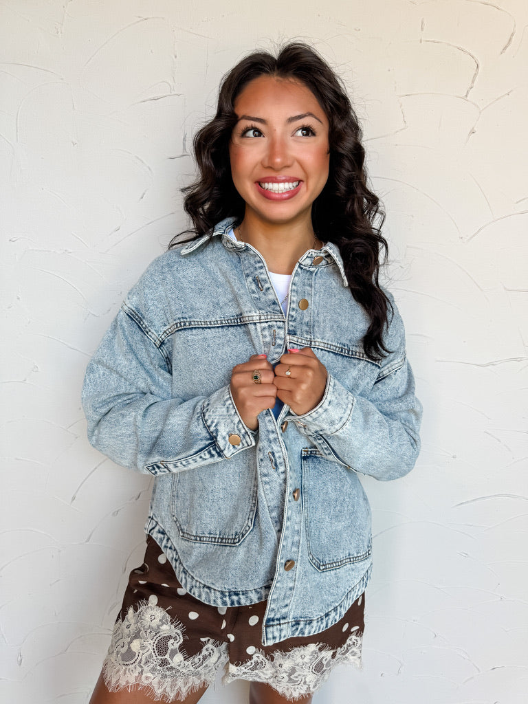A waist-up photograph of a smiling woman with long dark wavy hair looking away while holding the lapels of her jacket against a textured white wall. She is wearing an oversized light-wash denim jacket made from an 80% cotton and 20% polyester blend, featuring gold-tone button hardware, dropped shoulders, and front patch pockets, layered over a white top and paired with brown polka dot shorts with white lace trim.