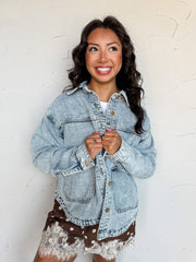 A waist-up photograph of a smiling woman with long dark wavy hair looking away while holding the lapels of her jacket against a textured white wall. She is wearing an oversized light-wash denim jacket made from an 80% cotton and 20% polyester blend, featuring gold-tone button hardware, dropped shoulders, and front patch pockets, layered over a white top and paired with brown polka dot shorts with white lace trim.