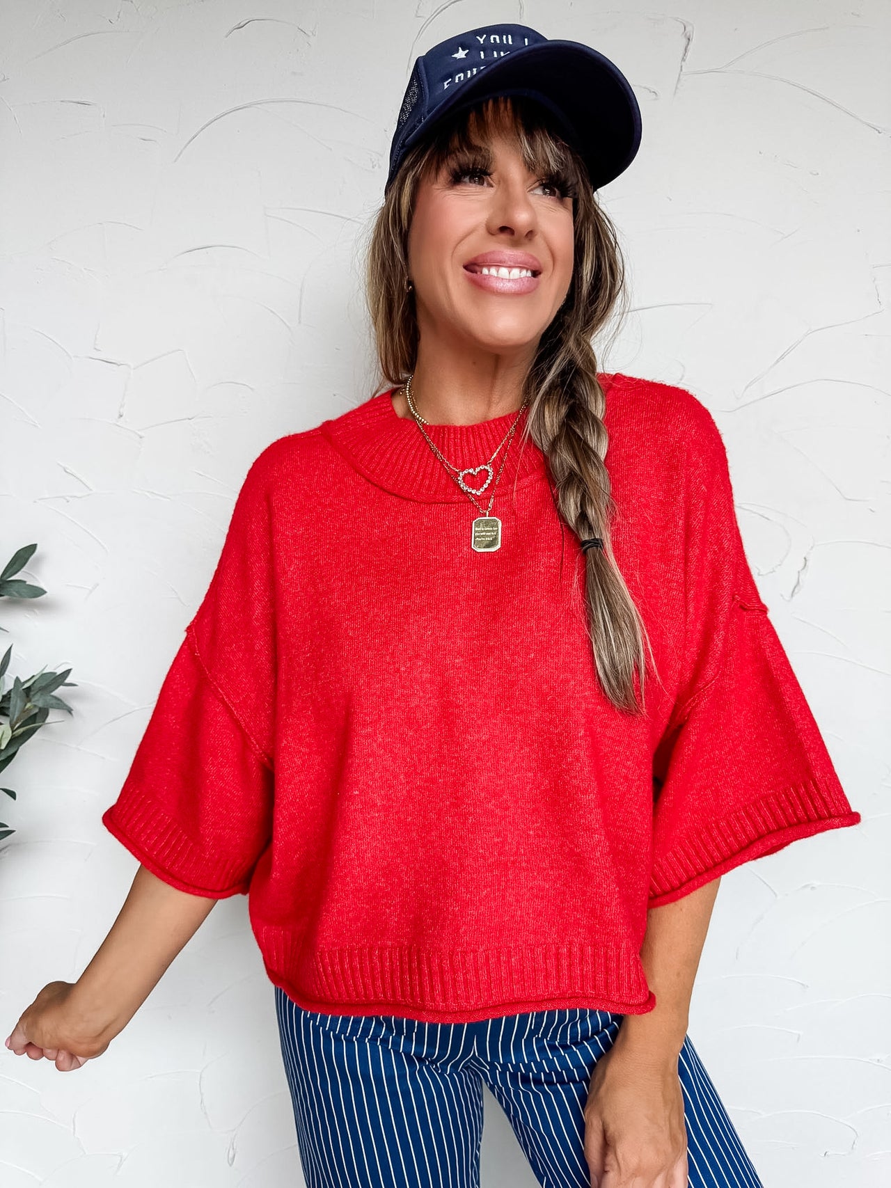 A woman smiling and looking to the side against a textured off-white wall, wearing a bright red knit sweater crafted from a blend of 65% nylon, 30% acrylic, and 5% wool. The top features a wide mock neckline, dramatic drop shoulders, and relaxed short sleeves finished with ribbed cuffs. The slouchy, oversized silhouette is accented by a coordinating ribbed hemline. She is styled with a navy blue trucker hat, layered gold necklaces including a heart charm and a square pendant, and navy blue and white pinstri