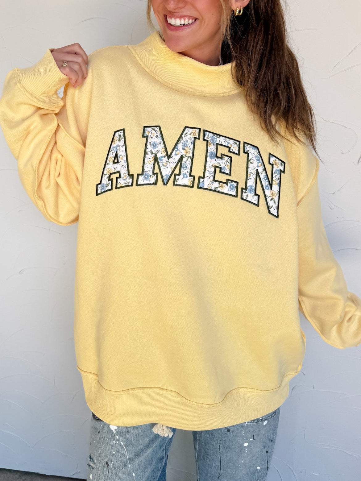 Person wearing a yellow sweatshirt with 'AMEN' printed on it against a white wall.