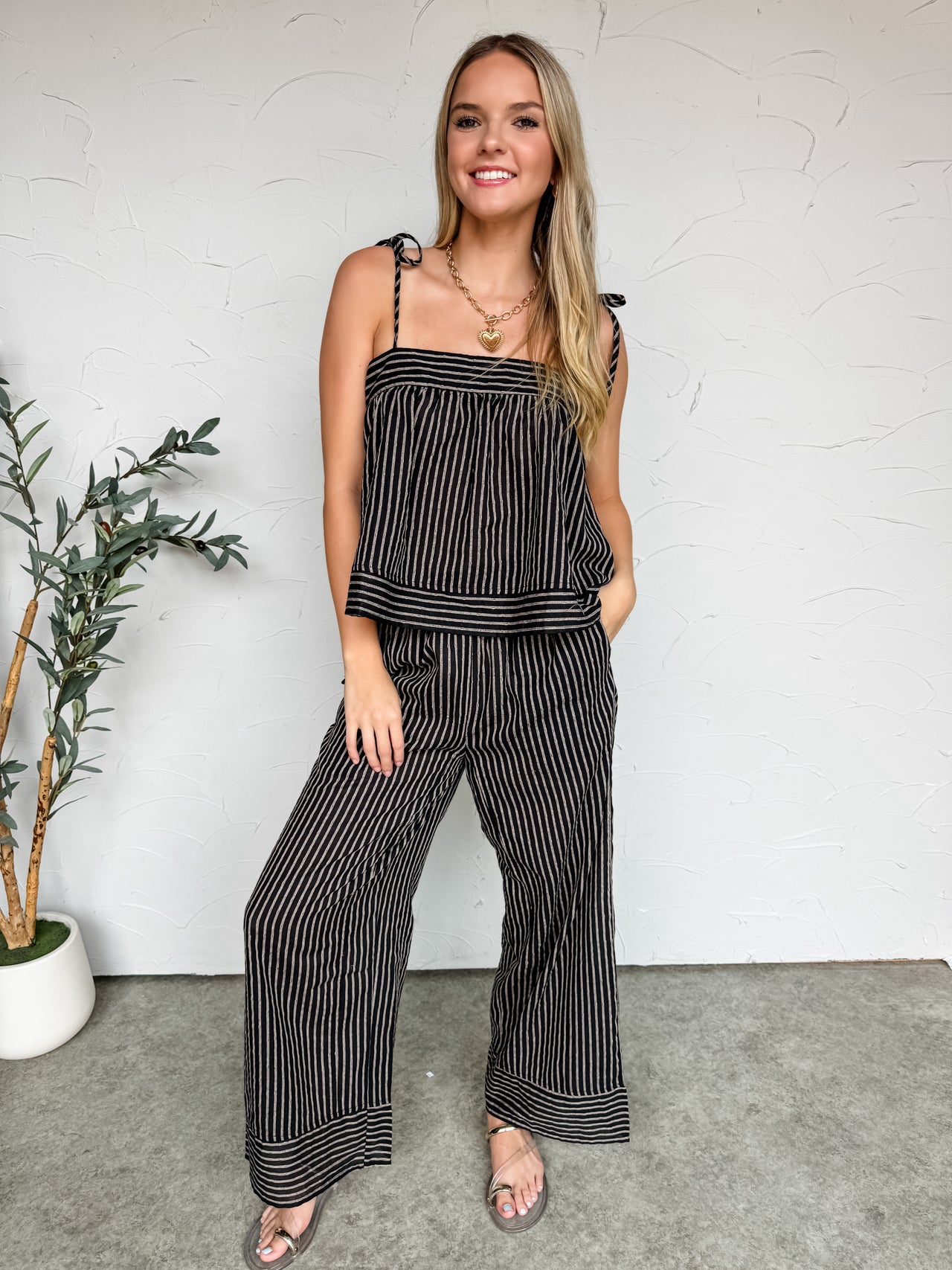 A full-length photo of a smiling blonde woman standing against a textured white wall, wearing a matching two-piece black and off-white pinstriped lounge set. The outfit features a flowy, loose camisole top with self-tie shoulder straps and relaxed, wide-leg trousers. She has one hand in her pocket and is accessorized with a gold chain heart necklace and metallic strappy sandals, with a potted olive plant visible to her left.