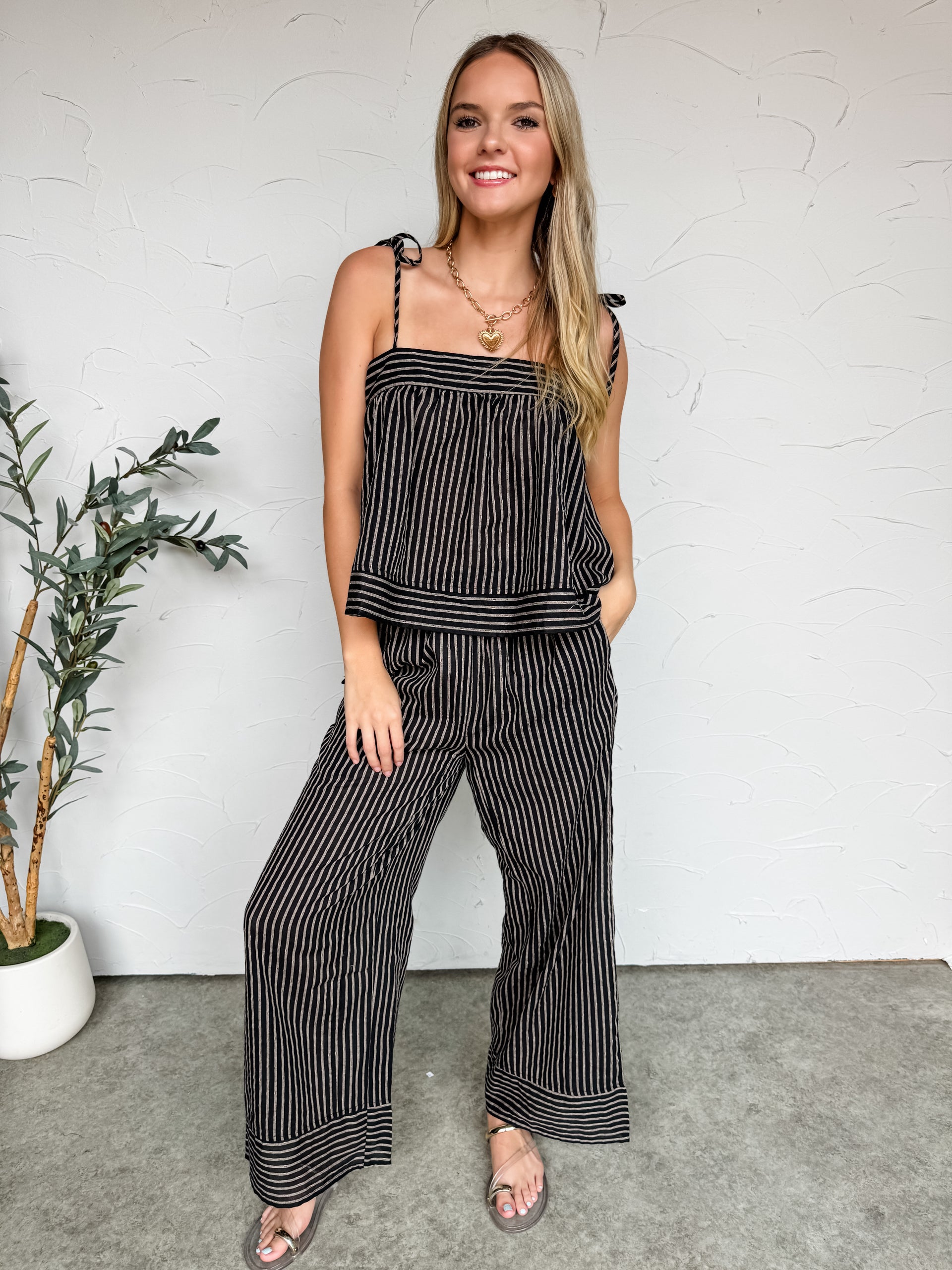 A full-length photo of a smiling blonde woman standing against a textured white wall, wearing a matching two-piece black and off-white pinstriped lounge set. The outfit features a flowy, loose camisole top with self-tie shoulder straps and relaxed, wide-leg trousers. She has one hand in her pocket and is accessorized with a gold chain heart necklace and metallic strappy sandals, with a potted olive plant visible to her left.