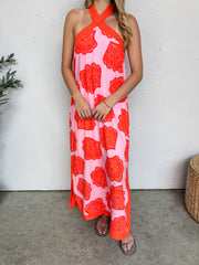 A woman smiling against a textured off-white wall, wearing a lined pink and orange maxi dress crafted from a 90% rayon and 10% linen blend fabric featuring an all-over patterned seashell print. The dress showcases a crossover halter neckline with solid vibrant orange crisscross straps that match the solid orange trim along the hemline and side slits. She is styled with a simple gold bracelet and metallic strappy flat sandals, standing on a concrete floor next to a potted faux olive plant and a woven wicker 