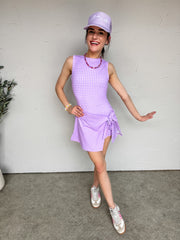 Person wearing a purple checkered dress and cap against a white wall.