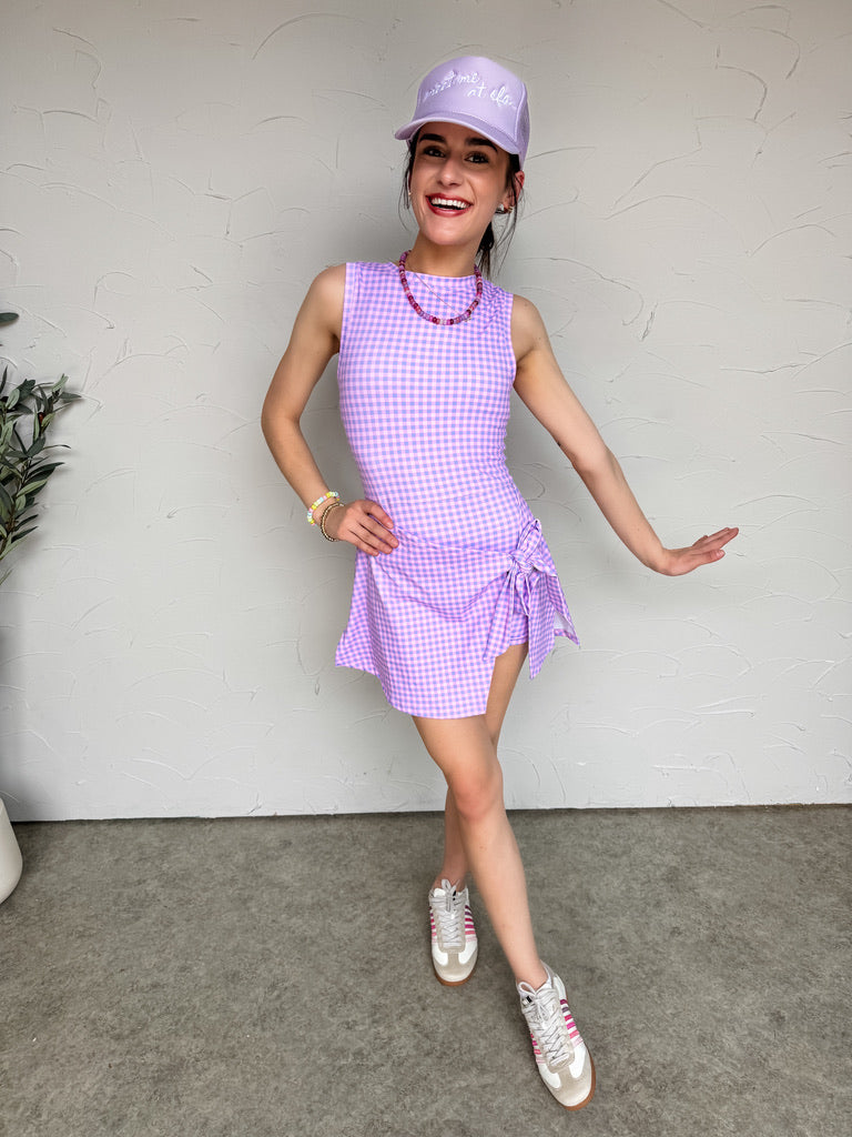 Person wearing a purple checkered dress and cap against a white wall.