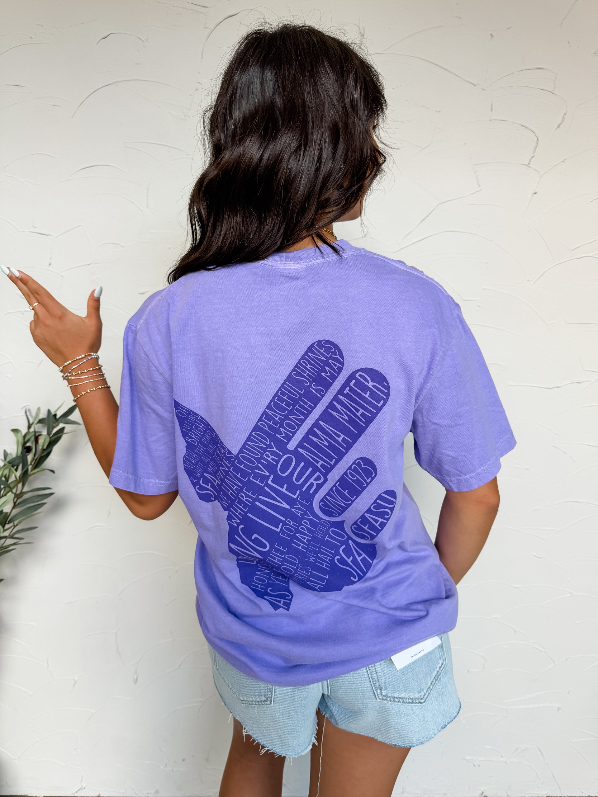 Person wearing a purple t-shirt with a blue graphic design on the back, standing against a white wall.