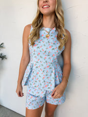 A close-up shot of a woman smiling and leaning against a textured off-white wall, wearing a matching two-piece set crafted from a 97% cotton and 3% spandex blend seersucker fabric. The light blue and white fine pinstripe set features an all-over coral-pink ditsy floral print. The sleeveless top is designed with a round neckline, fitted vertical princess seams, and a gathered peplum hemline, paired with matching high-waisted shorts. She is styled with a chunky gold link chain necklace featuring a textured he