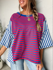 A woman laughing and posing with arms outstretched against a textured off-white wall, wearing an oversized colorblock knit t-shirt crafted from a 55% cotton and 45% polyester blend fabric. The top features mixed striped patterns, with the main front panel showing fine red and blue horizontal stripes with a solid blue crew neckline. The oversized drop-shoulder short sleeves and flared side panels feature a contrasting blue and white striped pattern. She is styled with a green camouflage trucker hat decorated