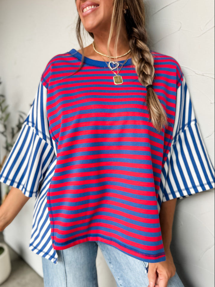 A woman laughing and posing with arms outstretched against a textured off-white wall, wearing an oversized colorblock knit t-shirt crafted from a 55% cotton and 45% polyester blend fabric. The top features mixed striped patterns, with the main front panel showing fine red and blue horizontal stripes with a solid blue crew neckline. The oversized drop-shoulder short sleeves and flared side panels feature a contrasting blue and white striped pattern. She is styled with a green camouflage trucker hat decorated